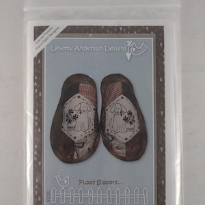 Może przedstawiać: Zestaw wzorów rzemieślniczych Lynette Anderson Designs zatytułowany Puppy Slippers, zawierający ilustrację dwóch kapci z grafiką szczeniaka i doniczki, naprasowanki oraz wstępnie wycięte papierowe sześciokąty w przezroczystej plastikowej osłonie.