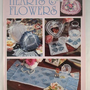 Può includere: Una copertina di libro rosa intitolata "HEARTS & FLOWERS" con immagini di progetti di uncinetto a filo. I progetti includono centrini, una decorazione a forma di cuore e un runner da tavolo. Il libro è di Anne Halliday.