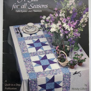 Puede incluir: Portada del libro "A Star for all Seasons" con un camino de mesa y manteles individuales. El camino de mesa tiene un patrón de estrellas en azul y blanco. Una taza de té, cubiertos y flores están sobre la mesa.