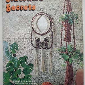 May include: A vintage craft book titled Macramé Secrets featuring a circular wall mirror with cream and brown knotted cord accents, a multi-tiered brown macramé plant hanger with green foliage, and text reading Exciting new designs.