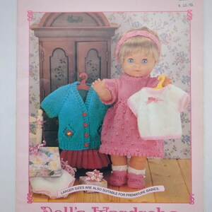 May include: A Sirdar 244 knitting pattern booklet titled Doll's Wardrobe, 13 Complete Outfits For Your Baby Doll. The cover features a doll wearing a pink knitted dress, headband, and booties, holding a white top, with a teal cardigan nearby.