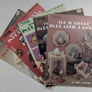 May include: A collection of six Leisure Arts craft leaflets arranged in a fan shape. The top leaflet is titled The Seashore in Plastic Canvas by Dick Martin, featuring images of tissue box covers and coasters decorated with seashells and sea creatures.