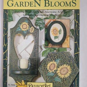 以下が含まれることがあります： 「Garden Blooms」というタイトルのクラフトブック。ひまわりのデザインが施された装飾的な木製アイテムを紹介しています。表紙には、ハート型のキャンドルホルダー、額入りのひまわり、六角形の作品があり、すべてに使い古されたような仕上がりになっています。