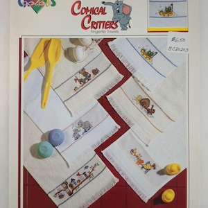 May include: A collection of white fingertip towels with embroidered cartoon animal designs. The towels feature various critters, including elephants, bears, and other whimsical characters. The towels are displayed on a red surface, with the "Comical Critters" logo visible.