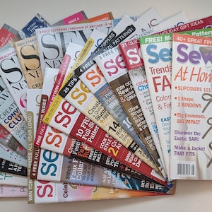 Può includere: Una collezione di riviste Sew News disposte a ventaglio. Le copertine presentano vari titoli di progetti di cucito, tra cui Slipcovers 101, 40+ Great Finds e 3 Free Patterns. Le riviste mostrano grafiche e testi colorati.