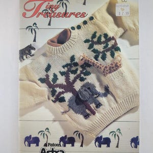 Può includere: Un libro di modelli di maglieria vintage intitolato "Tiny Treasures" di Patons. La copertina presenta un maglione lavorato a maglia color crema con un disegno di elefante e albero. Il maglione ha bottoni verdi e un giocattolo giraffa.