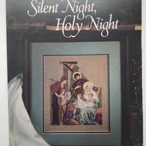 Puede incluir: Un folleto de manualidades de Leisure Arts titulado Silent Night, Holy Night por Carol Emmer. La portada muestra un patrón de punto de cruz de una escena de natividad con figuras en un establo, enmarcado en madera oscura con paspartú verde.