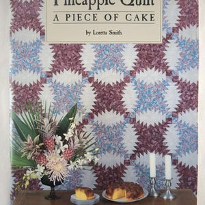 May include: The book cover for "Pineapple Quilt A Piece of Cake" by Loretta Smith. It shows a quilt pattern in purple, blue, and white. A table is set with a cake, a slice of cake, flowers, and candles.