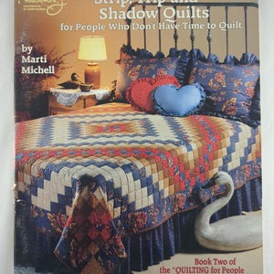 May include: A book cover titled "Strip, Trip and Shadow Quilts for People Who Don't Have Time to Quilt" by Marti Michell. The image shows a bed with a patchwork quilt in shades of blue, red, and brown, along with decorative pillows and a swan figurine.