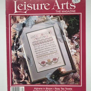 May include: A vintage issue of "Leisure Arts The Magazine" from April 1995, featuring a framed cross-stitch sampler. The cover is red with white text and includes a price tag. The sampler has floral and alphabet designs.