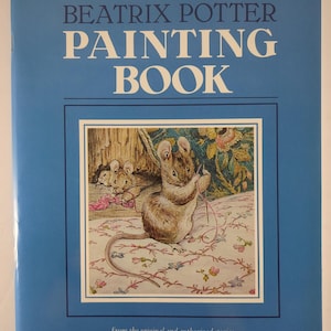Pode incluir: Um livro de capa mole azul intitulado The Big Beatrix Potter Painting Book. A capa apresenta uma ilustração central de um rato castanho a tricotar com fio cor-de-rosa numa superfície com padrão floral, com texto de Beatrix Potter e F. Warne & Co.