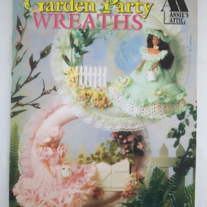 Puede incluir: Portada de revista "Fashion Doll Garden Party Wreaths" con dos muñecas con vestidos de ganchillo, sentadas en coronas en forma de media luna adornadas con flores y vegetación. El fondo es azul claro y blanco, con el texto "Annie's Attic".