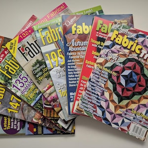 Peut inclure: Une collection de magazines Fabric Trends disposés en éventail, présentant des motifs de courtepointe colorés sur les couvertures. Les titres incluent Fabric Trends, avec du texte mettant en avant des créateurs et des techniques.