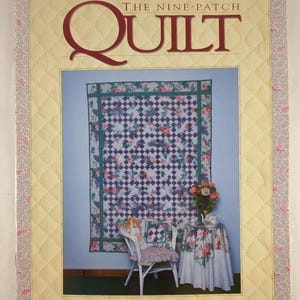 May include: A book cover titled "The Nine-Patch Quilt" with a floral quilt design. The cover has a quilted yellow border and a floral patterned border. A chair and table are set up in front of the quilt.
