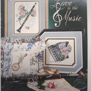 May include: A Stoney Creek Collection book 78 titled Love is the Music featuring cross-stitch patterns of a clarinet, a banjo on a ruffled floral pillow, and an accordion. A black clarinet and sheet music with a pink rose lie in the foreground.