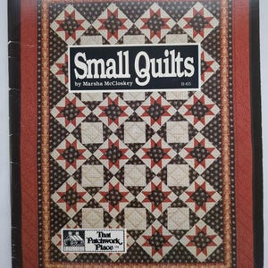 Puede incluir: Una portada de libro titulada "Small Quilts" de Marsha McCloskey. La portada presenta un diseño de edredón con estrellas y cuadrados rojos, marrones y crema. El libro tiene un borde rojo y un fondo negro.
