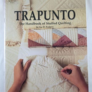 Puede incluir: Un libro titulado "Trapunto: The Handbook of Stuffed Quilting" de Sue H. Rodgers. La portada presenta una pila de tela acolchada en tonos crema, melocotón y burdeos. Se muestra una mano cosiendo una pieza acolchada blanca.