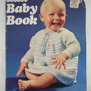 Peut inclure: Couverture de livre pour bébé vintage avec un bébé souriant vêtu d'une robe tricotée bleu clair et blanche avec des pompons. La couverture indique "PATONS Baby Book" et "No. 16". Le bas de la couverture indique "New collection of designs to knit and crochet".