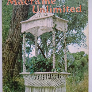 May include: A vintage magazine cover titled "Macrame Unlimited" features a macrame plant cradle. The plant cradle is white, with a fringed roof and a woven basket design. The magazine cover also includes the text "The Plant Cradle" and "New and Exciting Designs for Beginners and Pros."