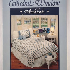 Könnte beinhalten: Buchcover mit dem Titel "Cathedral Window A Fresh Look" von Nancy J. Martin. Es zeigt ein Bett mit einer blau-weißen Steppdecke, Kissen und einem gerüschten Bettrock. Der Hintergrund zeigt ein Fenster mit Aussicht.
