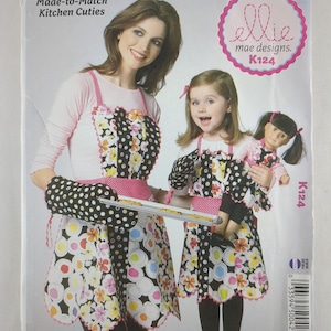 Pode incluir: Um padrão de costura "Made-to-Match Kitchen Cuties" com uma mulher e uma criança a usar aventais e luvas de forno a condizer. Os aventais têm um fundo preto com padrões florais coloridos e acabamentos cor-de-rosa. O padrão é da "ellie mae designs."