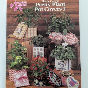 以下が含まれることがあります： プラスチックキャンバス製のプランターカバーを様々なデザインで紹介したクラフト本の表紙。本のタイトルは「Plastic Canvas Pretty Plant Pot Covers I」で、Annie's Atticから出版されています。表紙には、家、ハート、リボン、花など、様々なデザインのプランターカバーが描かれています。