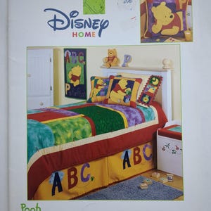 Op de afbeelding: Een Disney Home Peek-a-Boo Pooh Patchwork projectboek. De omslag toont een kleurrijk patchwork sprei met Winnie de Poeh-ontwerpen, ABC-letters en het Disney-logo. De afbeelding bevat een bed, kussens en een kleine kist.