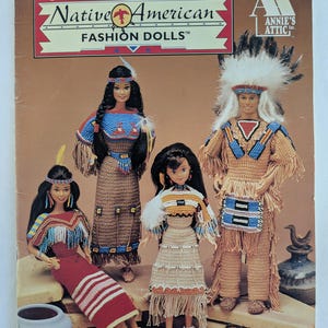 May include: A crochet pattern booklet for Native American fashion dolls. The cover features four dolls dressed in traditional attire, including a woman in a red dress with white stripes, a woman in a brown dress with fringe, a woman in a tan dress with fringe, and a man in a brown and blue outfit with a headdress. The booklet is titled "Native American Fashion Dolls" and is published by Annie's Attic.