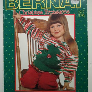以下が含まれることがあります： 緑色の表紙に白い模様が描かれたBernat Christmas Treasuresのクラフトブック。表紙には、赤、白、緑のストライプのニットセーターを着た子供が写っており、セーターには緑の木のデザインと小さなクリスマスのボタンがあしらわれています。