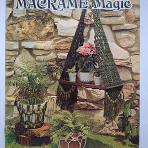 Peut inclure: Une couverture de magazine vintage intitulée "MACRAME Magic" présentant des suspensions et des jardinières en macramé. L'image présente une variété de plantes dans des supports en macramé sur un fond de mur en pierre. La couverture comprend une étagère en macramé avec des plantes en pot.