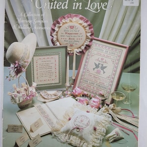 Puede incluir: Una colección de bordados de matrimonio y accesorios de boda. La portada presenta el título "United in Love" con varios diseños de punto de cruz, un sombrero decorativo, una cesta y tarjetas de mesa. La paleta de colores incluye rosa, blanco y verde.
