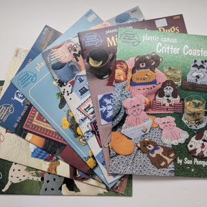 Op de afbeelding: Een verzameling American School of Needlework plastic canvas patroonboekjes in een waaier gerangschikt. Het bovenste boekje is getiteld Plastic Canvas Critter Coasters door Sue Penrod en bevat illustraties van onderzetters in diervorm.