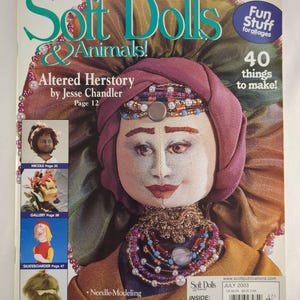 Puede incluir: La portada de la revista "Soft Dolls & Animals!" presenta una muñeca detallada con un tocado y collar enjoyados. La revista incluye artículos sobre modelado con aguja, teñido de telas y patrones de animales realistas. Se muestra el número de julio de 2003.