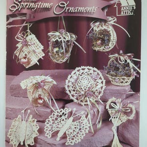 Puede incluir: Una colección de adornos de ganchillo de primavera en varias formas, incluyendo mariposas, corazones y cestas colgantes. Los adornos son de color crema con acentos rosas y morados. La imagen incluye el texto "Springtime Ornaments" y "Annie's Attic".