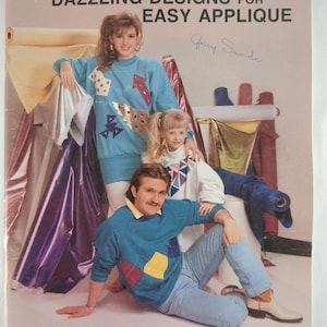 May include: A vintage sewing pattern cover titled "Dazzling Designs for Easy Applique" by Country Thread Designs. The cover features a family wearing applique sweaters in shades of blue, white, and yellow. The author's name is Ginger Pahmeier.