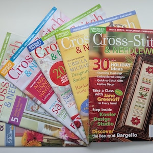 Pode incluir: Uma coleção de seis revistas Cross-Stitch & Needlework dispostas em leque. As capas apresentam várias cores, incluindo vermelho, amarelo e branco, com títulos em negrito e manchetes sobre ideias festivas, designs e artistas de bordado.