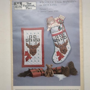 Peut inclure: Un patron de couture intitulé Reindeer Wall Hanging or Stocking par Christal Carter, 1986. La pochette présente une tenture murale rectangulaire et une chaussette de Noël, avec un motif de renne brun, des nœuds rouges et des dimensions de 47 cm par 76 cm.