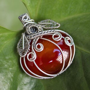May include: A wire-wrapped pendant featuring a smooth, rounded, deep orange carnelian stone encased in silver-toned metal wire. The wire is intricately coiled to form a stem and two leaf-like shapes at the top of the stone.