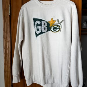 Könnte beinhalten: Ein cremefarbener Pullover mit einer grünen, gelben und weißen Applikation einer Flagge mit den Buchstaben "GB" und einem grünen Stern.