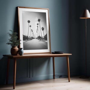 May include: A framed black and white photograph of a palm tree-lined street. The photograph is displayed on a wooden table against a dark blue wall. A floor lamp and decorative items are also present.