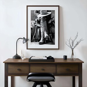 May include: Black and white framed print of a cowboy on a horse, wearing a hat and boots. The image is displayed on a wooden desk with a black lamp, candles, and a decorative tray.