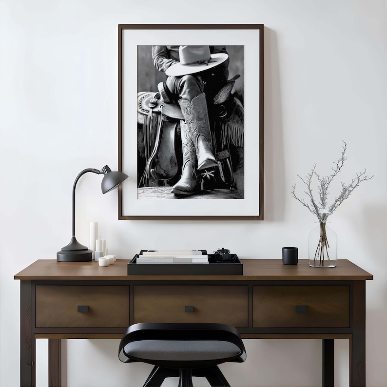 May include: Black and white framed print of a cowboy on a horse, wearing a hat and boots. The image is displayed on a wooden desk with a black lamp, candles, and a decorative tray.