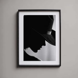 Puede incluir: Impresión enmarcada en blanco y negro que muestra un perfil de una persona con un sombrero de ala ancha con una banda oscura. La imagen utiliza la luz y la sombra para crear un efecto dramático.