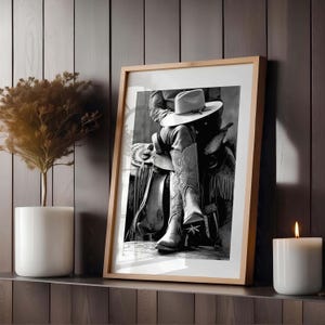 May include: Framed black and white photograph of a cowboy on a saddle, wearing a hat and cowboy boots. The boots have decorative stitching. The frame is light brown, and the image is displayed on a shelf with a candle and a plant.