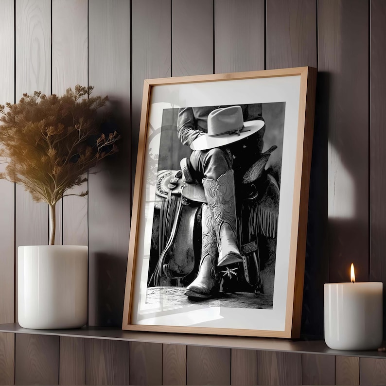 May include: Framed black and white photograph of a cowboy on a saddle, wearing a hat and cowboy boots. The boots have decorative stitching. The frame is light brown, and the image is displayed on a shelf with a candle and a plant.