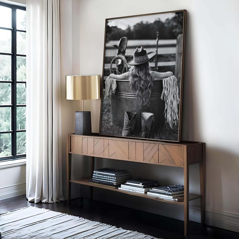 May include: A framed monochrome photograph of a person wearing a cowboy hat, seated in a metal tub, with cowboy boots in front. The photograph is on a wooden console table with a gold lamp.