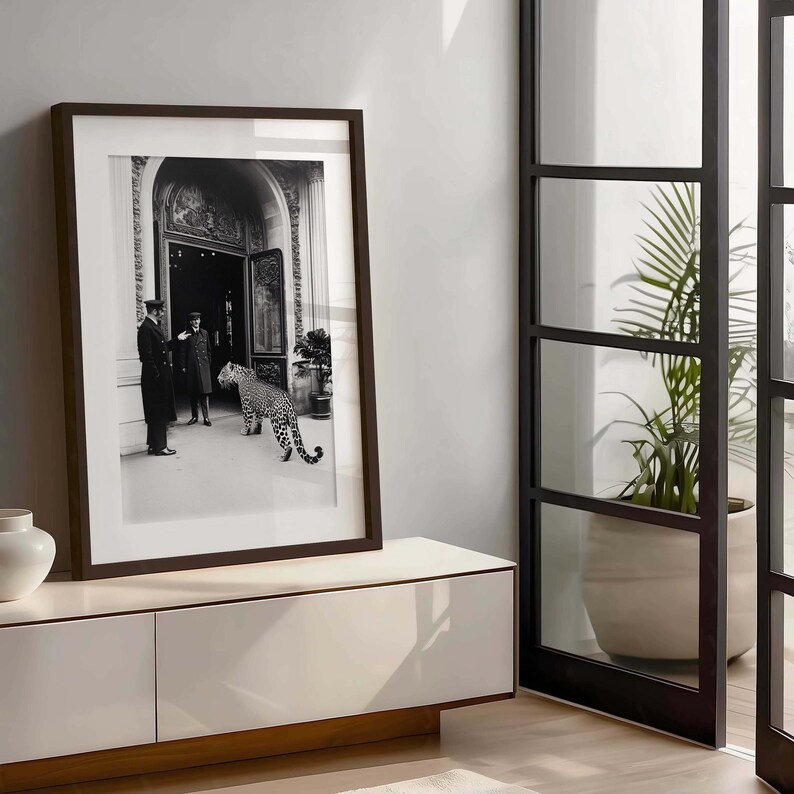 Op de afbeelding: Een zwart-witfoto van twee mannen in uniform die buiten een gebouw staan met een luipaard dat langs hen loopt. De foto is ingelijst in een donkerhouten lijst en hangt aan een witte muur.