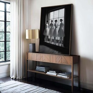May include: A framed black and white photograph of four ballerinas in white tutus standing by a window. The photograph is displayed on a wooden console table with a gold lamp and books. The room has a modern, minimalist aesthetic.