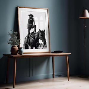 May include: Black and white framed print of a person in a cowboy hat and boots, standing on a horse. The print is displayed on a wooden table with a small plant and vase. The room has a dark blue wall and a floor lamp.