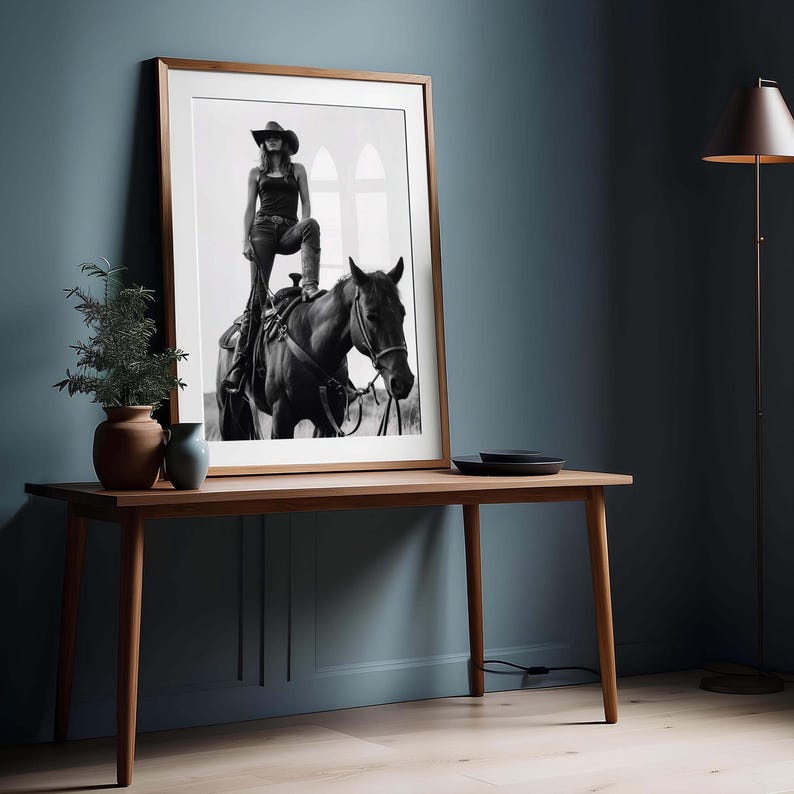 May include: Black and white framed print of a person in a cowboy hat and boots, standing on a horse. The print is displayed on a wooden table with a small plant and vase. The room has a dark blue wall and a floor lamp.